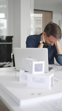 Male architect in glasses at a contemporary office working on a white architectural scale model of a...