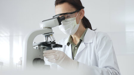 Female scientist wearing a lab coat, face mask, and safety glasses carefully adjusts a microscope,...