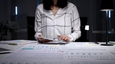 Businesswoman working late night in a modern office, analyzing financial data on a digital tablet...