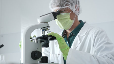 Medical scientist wearing protective mask, cap and green gloves using microscope doing scientific...