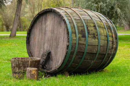 Large vintage wooden wine barrel with green metal bands placed on green grass in a park on a sunny day. Concept of traditional winemaking, rustic decor, and outdoor displayの写真素材