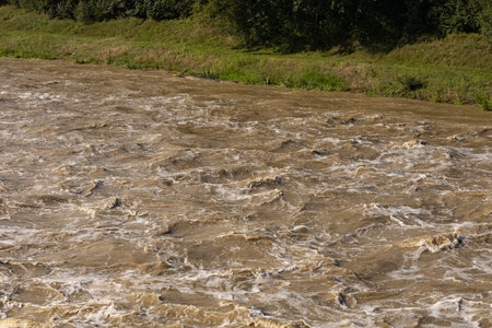 Powerful churning water near the riverbank with a strong current caused by heavy rainfall. The turbulent muddy water flows rapidly, creating intense waves demonstrating the force of natureの写真素材