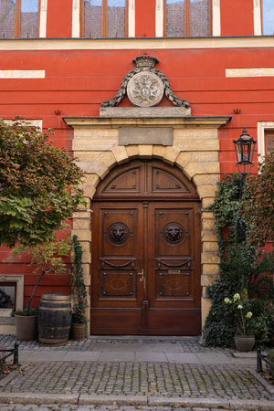 Elegant wooden Door with intricate carvings and decorative crest on historic red building, framed by vintage lanterns, ivy, and flowering plants, creating a charming European architectural sceneの写真素材