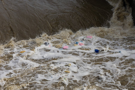 Polluted river water with foam and various plastic debris floating, bottles, and waste materials,...