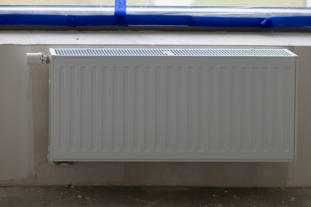Modern white panel radiator installed beneath a sealed window with blue protective tape during home renovation. Heating system installation, energy efficiency, and interior design upgradesの写真素材