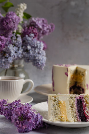 Elegant composition with a slice of poppy seed and berry cake on a white plate, lilac flowers, and a cup of tea, captured in soft natural light, evoking springtime freshness and cozy atmosphereの写真素材