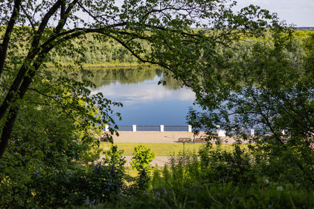 Scenic view of a tranquil river surrounded by lush green trees and a riverside promenade with benches on a sunny day. Relaxation, natural beauty and peaceful outdoor recreationの写真素材