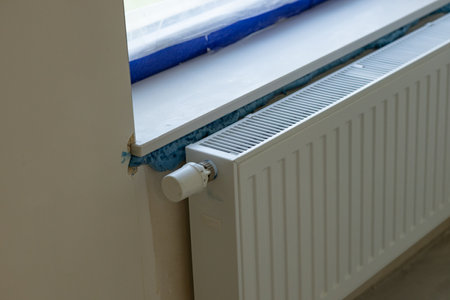 White home heating radiator with adjustable thermostat installed beneath a windowsill during renovation. Thermal insulation foam application, construction details and wall finishingの写真素材