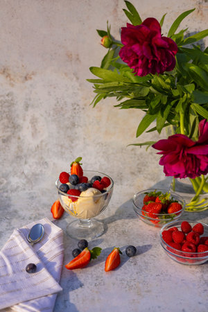 Elegant summer dessert of vanilla ice cream with fresh berries, served in a glass bowl on a textured background next to a bouquet of red peonies. Summer treats and floral decorの写真素材