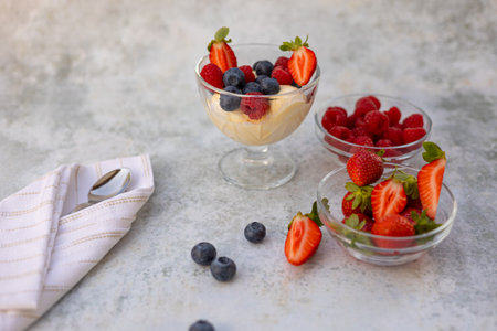 Refreshing vanilla ice cream dessert served with fresh berries in a glass bowl, surrounded by berry-filled dishes, a folded napkin, and a spoon on a textured light surfaceの写真素材