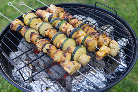 Grilled chicken and zucchini skewers cooking on a charcoal barbecue with baked potatoes wrapped in foil. Outdoor cooking, summer picnic and delicious barbecue food in a backyard settingの写真素材