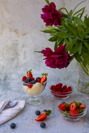 Elegant dessert composition with fresh berries and ice cream in glass bowls, accompanied by a bouquet of deep magenta peonies in a vase, creating a vibrant and stylish scene on a textured backgroundの写真素材