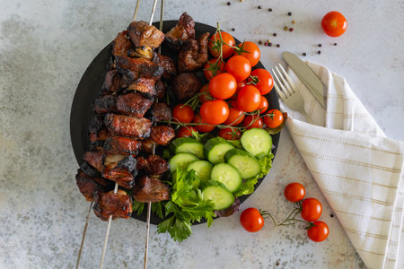 Top view of grilled meat skewers with fresh cherry tomatoes, cucumber slices, and parsley on a black plate, accompanied by cutlery wrapped in a napkinの写真素材