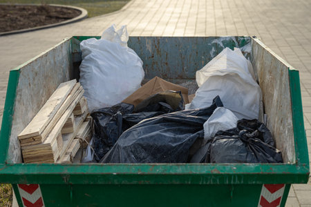 Close-up of a green industrial dumpster filled with construction debris, including wooden planks, black and white plastic garbage bags, and paper waste, placed outdoorsの写真素材
