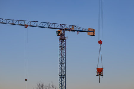 Close-up of a tower crane with a suspended orange concrete bucket against a clear blue sky during sunset. Construction technology, industrial equipment and modern architecture in progressの写真素材