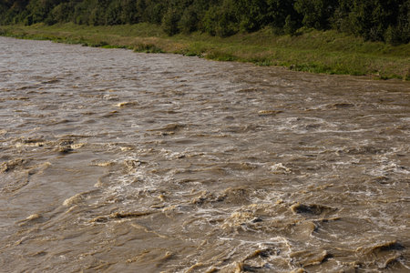 Fast-flowing muddy river with turbulent water caused by heavy rainfall passing through green forested riverbank on a sunny day. Flooding, extreme weather and natural disasterの写真素材
