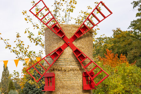 Artistic windmill made of straw bales with bright red wooden blades, set in a vibrant autumn scene. Surrounded by colorful trees and festive orange flagsの写真素材