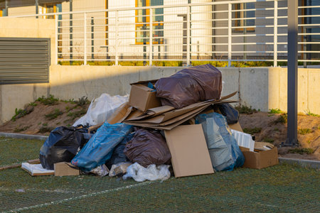 Pile of mixed garbage with black and blue plastic bags, cardboard boxes dumped near a modern residential area. Urban pollution, waste disposal, and environmental issuesの写真素材