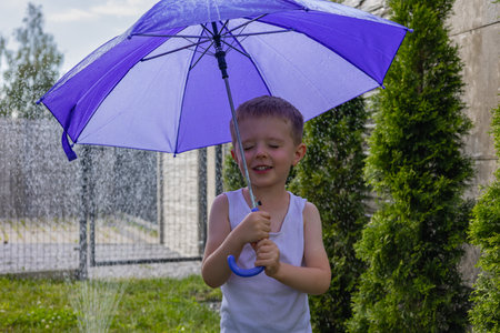 Happy boy with a playful smile holding a purple umbrella in the backyard during summer, standing under a garden sprinkler. Childhood joy, water games and outdoor fun on a sunny dayの写真素材