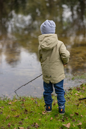 Little child in warm autumn clothes standing on green grass near a calm lake, holding a stick and looking at the water. Childhood curiosity, outdoor adventure, exploration and nature connectionの写真素材
