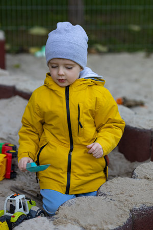Little boy in a yellow jacket and gray beanie playing in a sandbox with toy trucks and a plastic shovel on a cool autumn day. Childhood, outdoor play, creativity, imagination and early developmentの写真素材