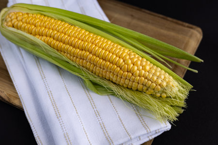 Fresh ear of corn with vibrant golden kernels partially wrapped in green husk placed on a wooden cutting board with a white linen napkin. Organic food, healthy eating and farm-to-table produceの写真素材