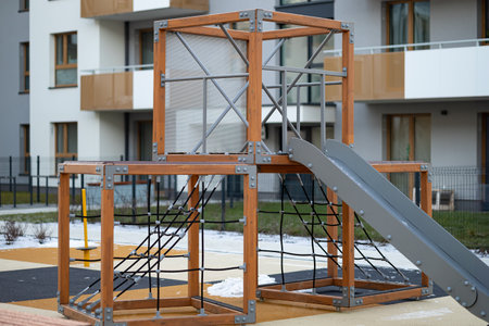 Modern playground of children with wooden climbing frames, rope nets, and a metal slide, located in a residential area with apartment buildings. Safe outdoor activities and urban family lifeの写真素材