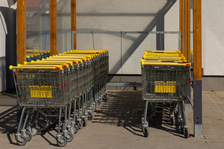 Multiple metal shopping carts with yellow handles neatly arranged in designated outdoor area at supermarket entrance on sunny day. Retail, consumerism shopping infrastructure and commerceの写真素材