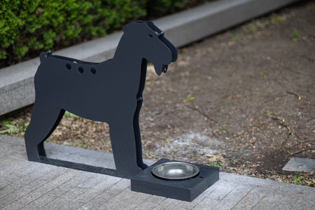 Metal dog-shaped water station with stainless steel bowl on city sidewalk, providing hydration for pets during hot summer days. Animal care, urban pet amenities and responsible ownershipの写真素材
