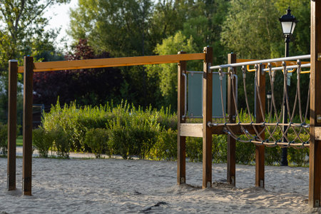 Empty outdoor playground with wooden rope bridge and horizontal bar on sandy ground in green park....