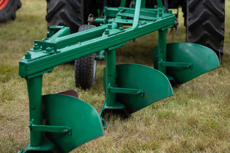 Green agricultural plow attached to tractor standing on grassy field, close up of metal blades and...