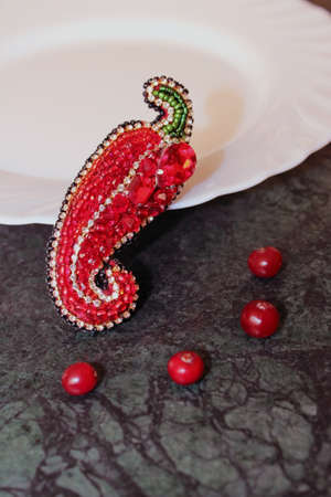A beaded brooch lies on a green marble table in front of an empty white plate. Brooch in the shape of a red bell pepper. On the table are red cranberries. Diet.の写真素材