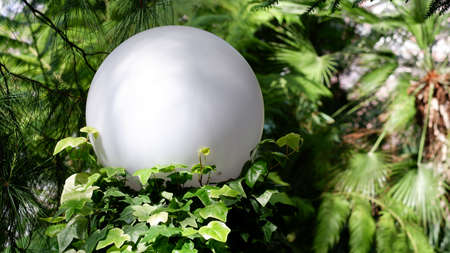 A white spherical lantern on an iron pole lighting a garden against a background of green plants in a southern city. Background from tropical plants. Copy space on a white background of a lantern.の写真素材