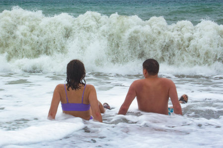 Rear view of a young couple sitting on the shore in sea water. There is sea foam around the girl and the boy. Strong waves crash on the shore. People are resting on the seashore.の写真素材