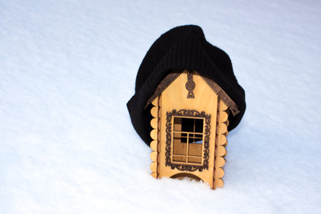 A wooden toy house stands in the snow in winter. The house is covered with a warm hat. The concept of keeping warm.の写真素材