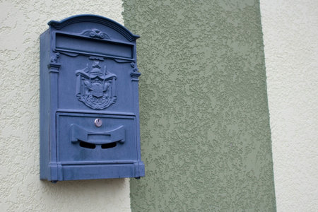 A gray mailbox on the wall of a building on one of the city streets. The box is locked with a key.
