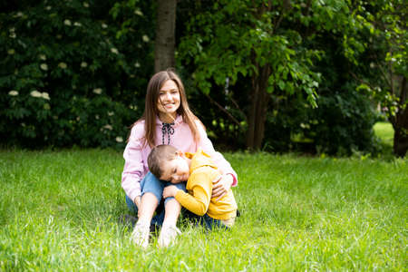 Young woman mom sits on green grass in summer park with boy son. Son hugs his mom.の写真素材