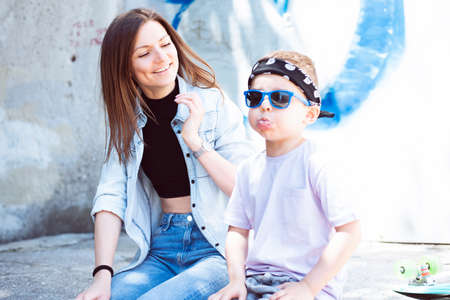 A young beautiful woman in a black sports top and a blue denim shirt sits with her boy-son in stylish clothes against the background of a concrete wall with graffiti.の写真素材