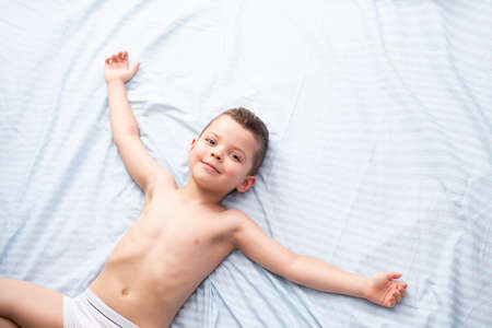 A little five-year-old boy in white underwear lies relaxed on a light blue bed and smiles.の写真素材