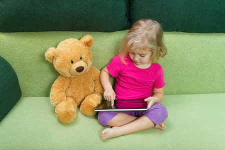 Little girl with Teddy bear, and a tablet. Sitting on a green couch.の写真素材