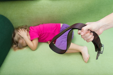 Family violence and aggression concept - furious angry man raised punishment hand holding leather belt over scared or terrified childの写真素材