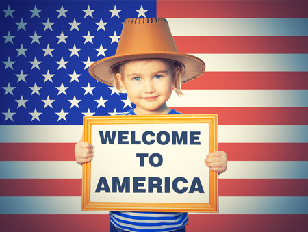 Little Funny girl in striped shirt with blackboard. Text welcome to America. On background of American flag.の写真素材
