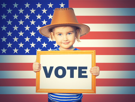 Little Funny girl in striped shirt with blackboard. Text VOTE. On background of American flag.の写真素材