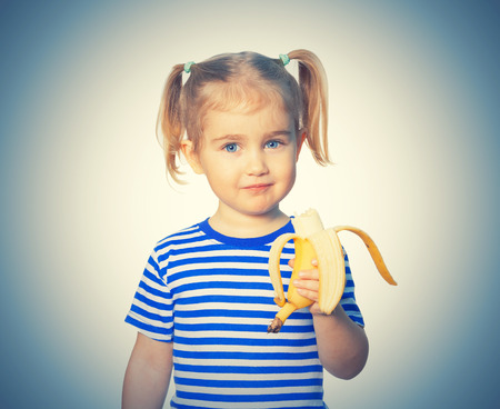Little Funny girl in striped shirt eats banana. Isolated on white backgroundの写真素材