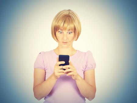 Closeup portrait anxious  young woman looking at phone seeing bad news. Blonde girl in horror reading sms on your smartphone isolated on  background. の写真素材
