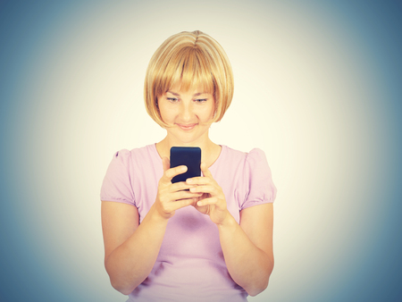 Young woman reading good news on the phone. Girl smiling blond looking at smartphone. Pleasant SMS. Isolated on background. の写真素材