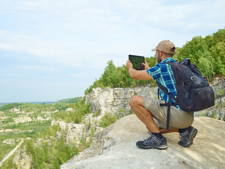 Man Tourist Uses Tablet Computer Sitting on Edge of a Cliff In Mountains.の写真素材