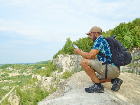 Man Tourist Uses Tablet Computer Sitting on Edge of a Cliff In Mountains.の写真素材