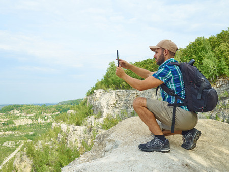 Man Tourist Uses Tablet Computer Sitting on Edge of a Cliff In Mountains.の写真素材
