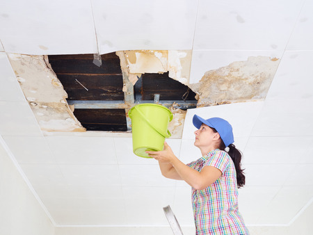 Young Woman Collecting Water In Bucket From Ceiling. Ceiling panels damaged huge hole in roof from rainwater leakage.Water damaged ceiling .の写真素材